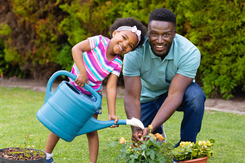 A smiling man and young girl are gardening, with the girl holding a blue watering can over blooming flowers - https://www.richardgreeninsurance.com/