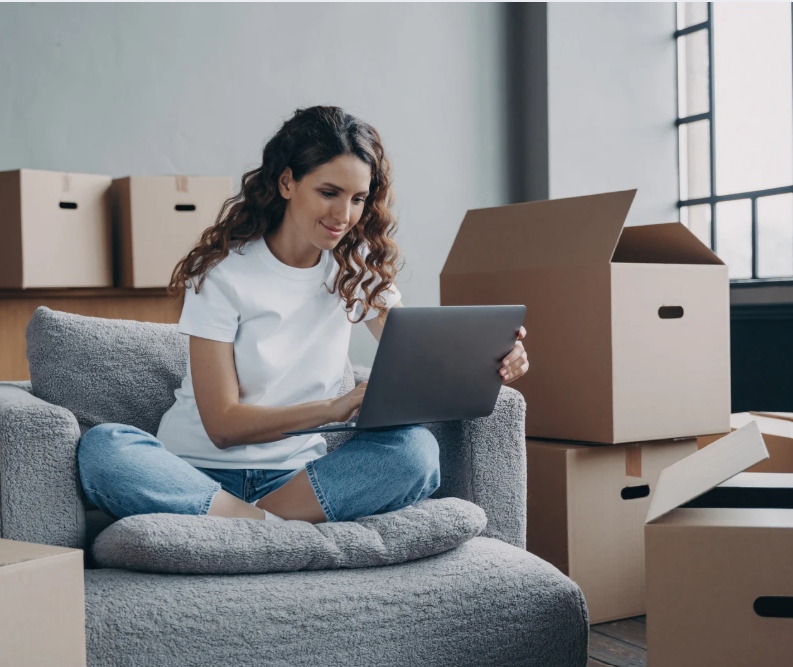 A smiling young woman sits cross-legged on a couch, surrounded by numerous cardboard moving boxes, using a laptop